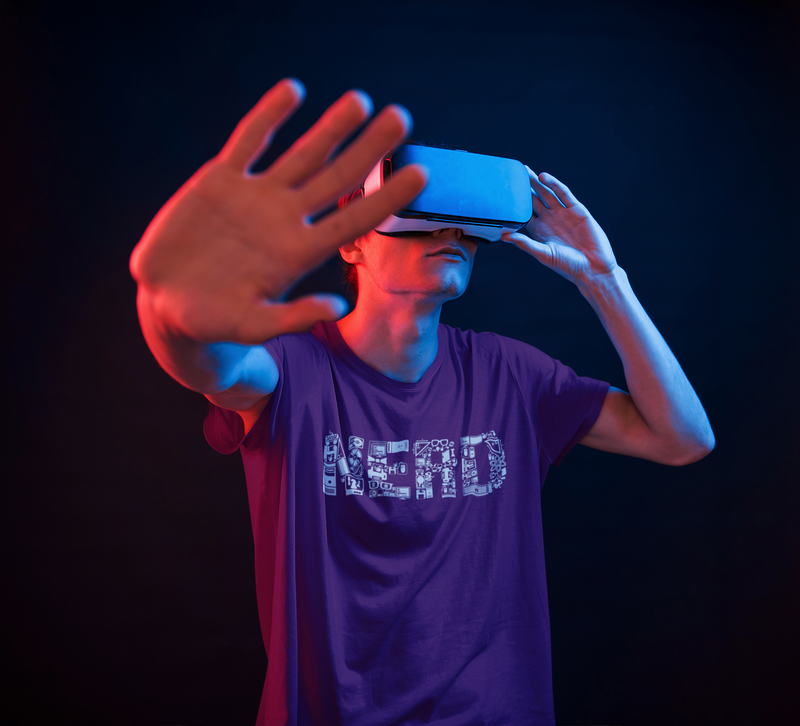 Guy wearing purple short sleeve t-shirt with an all white print of science and technology objects making the shape of the word "Nerd".